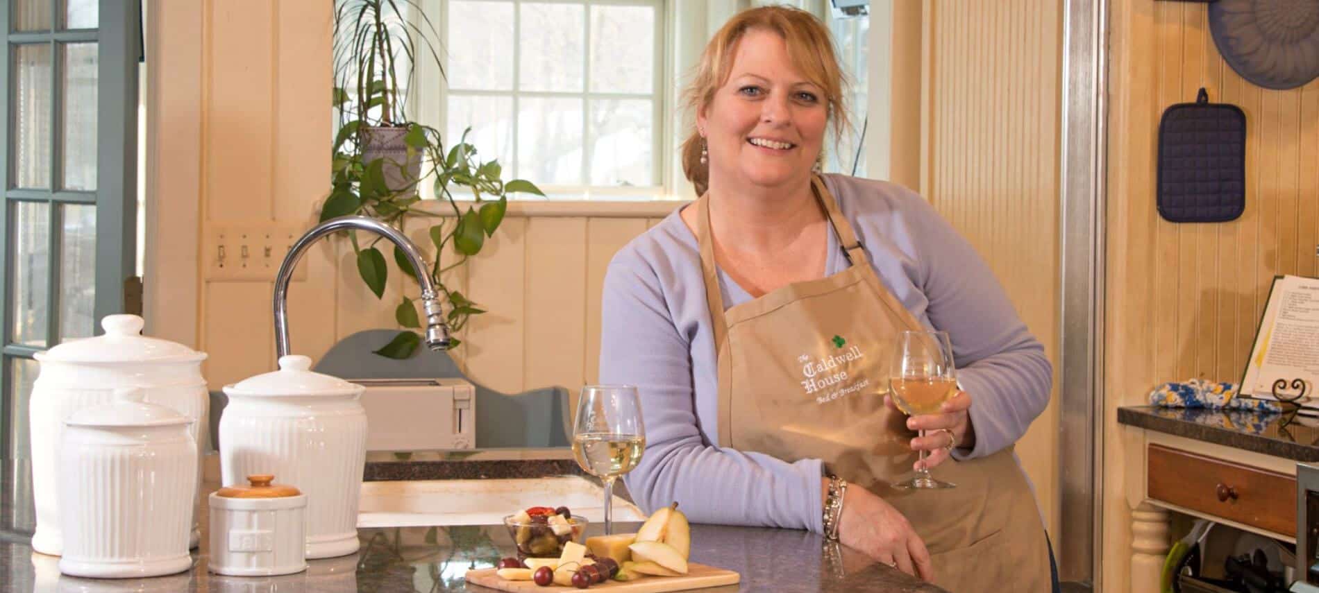 A proud and happy innkeeper smiles as she displays her charming kitchen, a selection of fine foods and wine